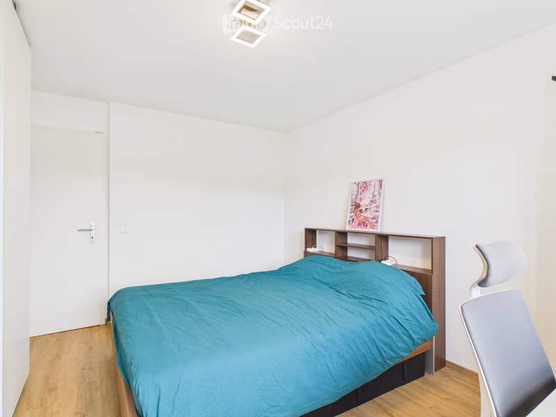 A modern, minimalist bedroom with a single bed covered in a teal duvet, a wooden shelf with a picture frame, a white chair, and a white door.