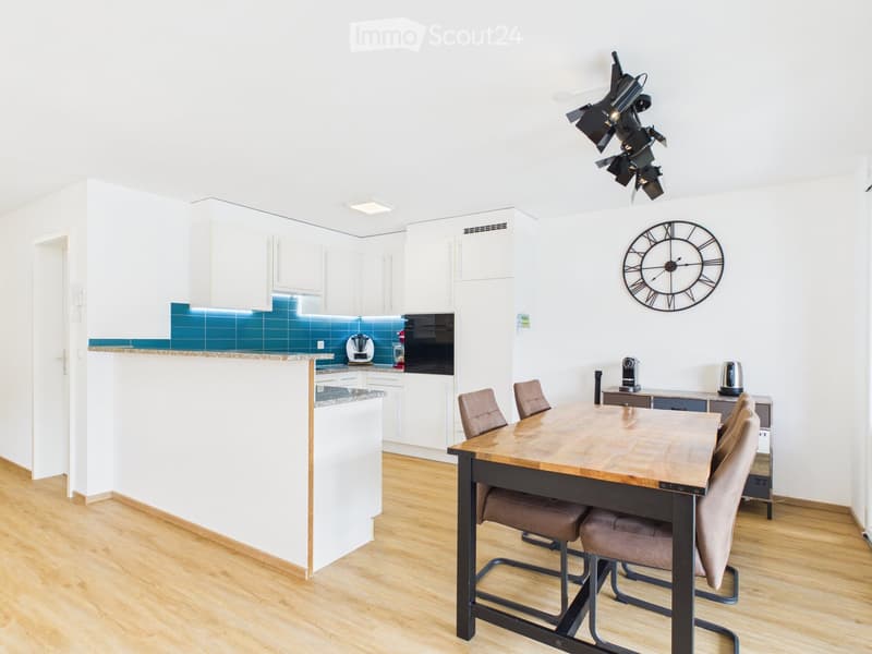 Modern kitchen and dining area with white cabinets, wooden flooring, a large wooden dining table with four chairs, and various appliances including a refrigerator, oven, and coffee maker.