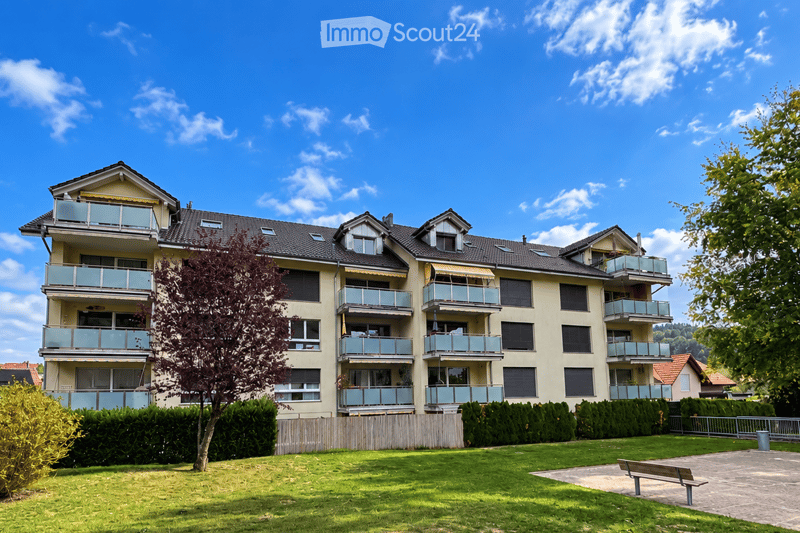 Multi-story residential building with multiple balconies, surrounded by greenery and a small park area with a bench.