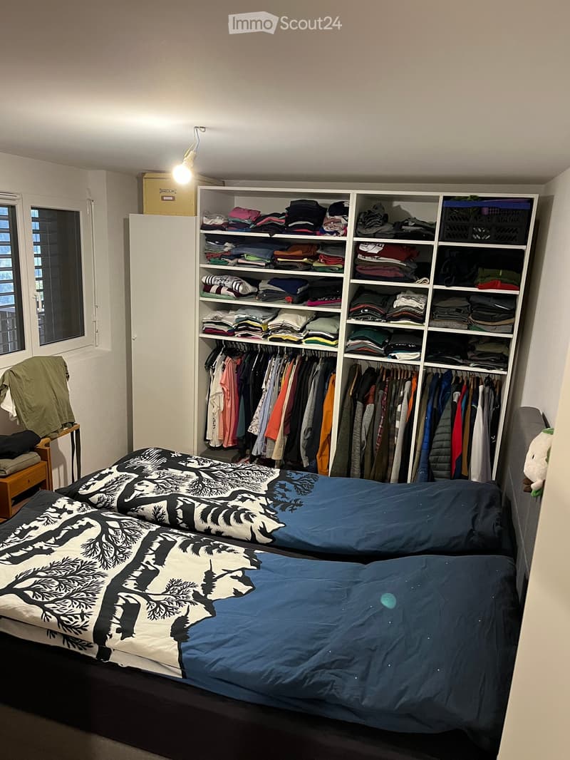 A tidy bedroom with a double bed, neatly made with blue and black bedding, a large wardrobe filled with folded clothes and hanging garments, and a small wooden nightstand with folded blankets and a green jacket draped over it.