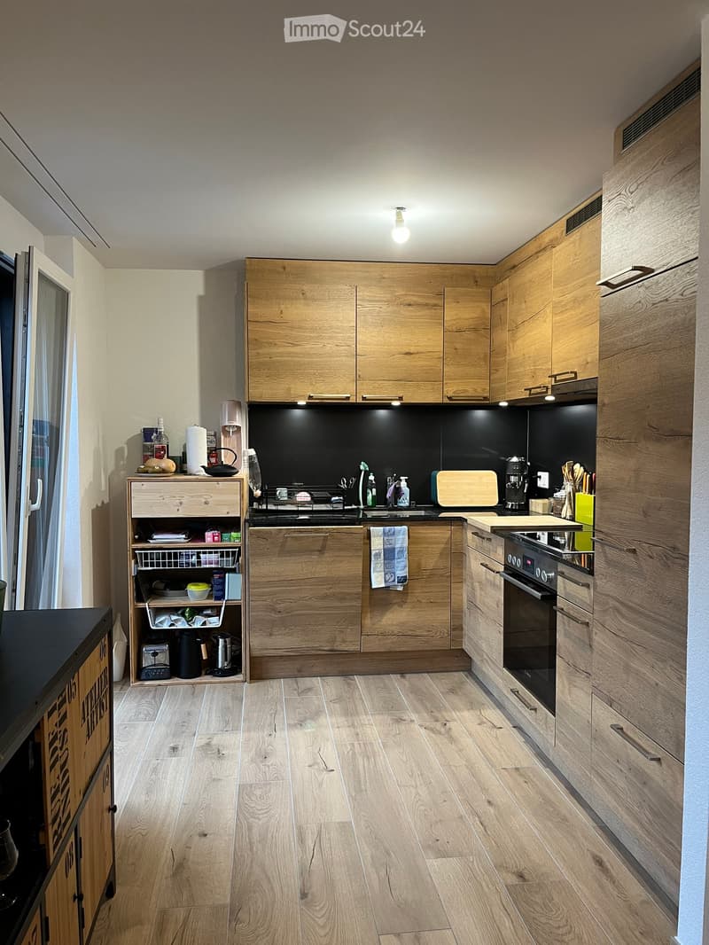 Modern kitchen with wooden cabinets, stainless steel appliances, and a black backsplash. Features include an oven, refrigerator, sink, and various small appliances on the counter.
