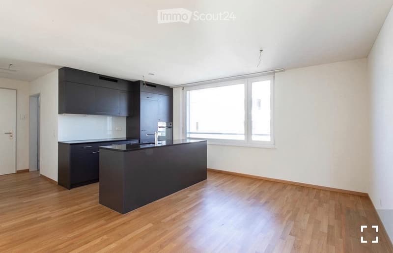 Modern kitchen with dark cabinets, a central island, and a large window providing natural light. The floor is made of light wood.