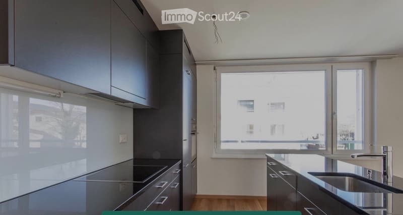 Modern kitchen with dark grey cabinets, stainless steel sink, and sleek countertops. Large windows provide natural light.