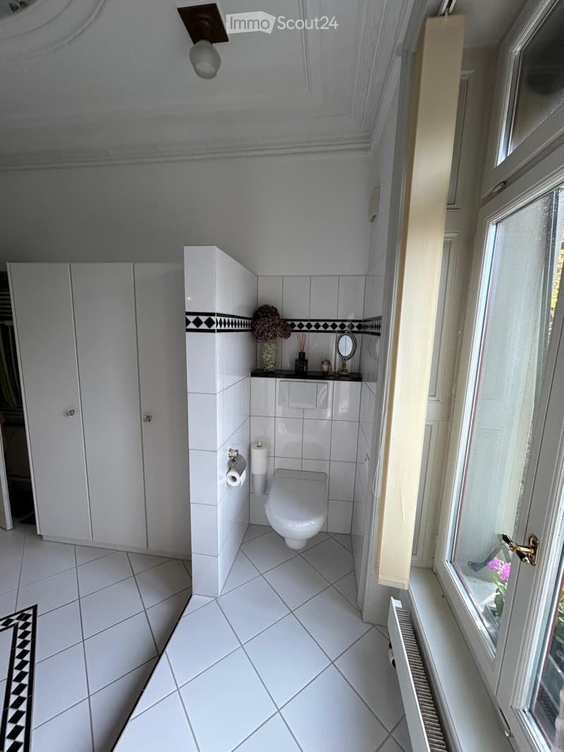 A small, modern bathroom with white tiles, a toilet, a mirror, a cabinet, and a vase with flowers on the counter.