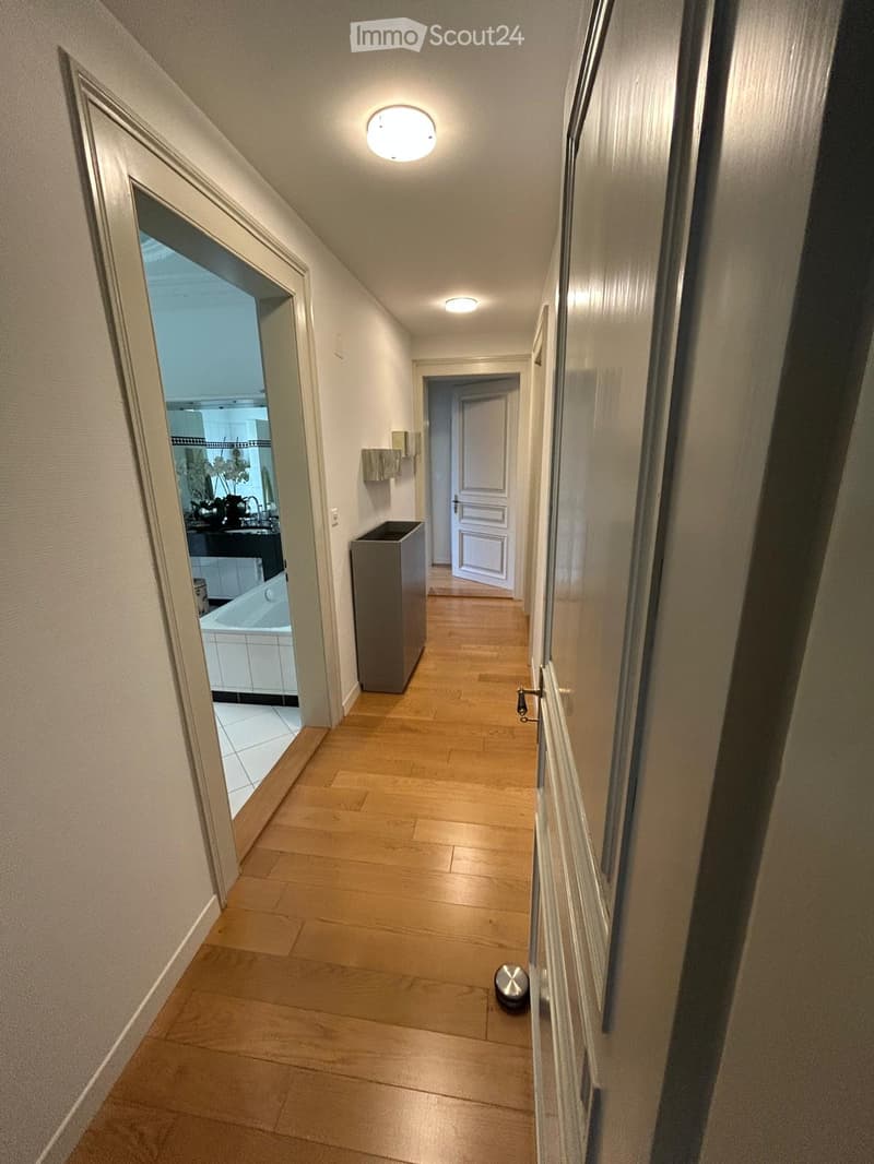 A narrow hallway with wooden flooring, white walls, and recessed lighting. An open door on the left leads to a bathroom with a bathtub and sink, while a closed door on the right leads to another room.