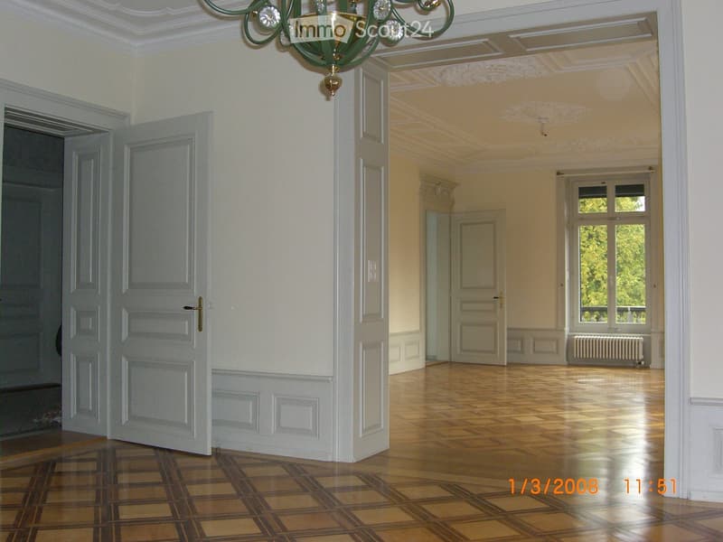 Empty hallway with wooden flooring, white walls, and a chandelier hanging from the ceiling. The hallway leads to another room with large windows and a radiator visible.