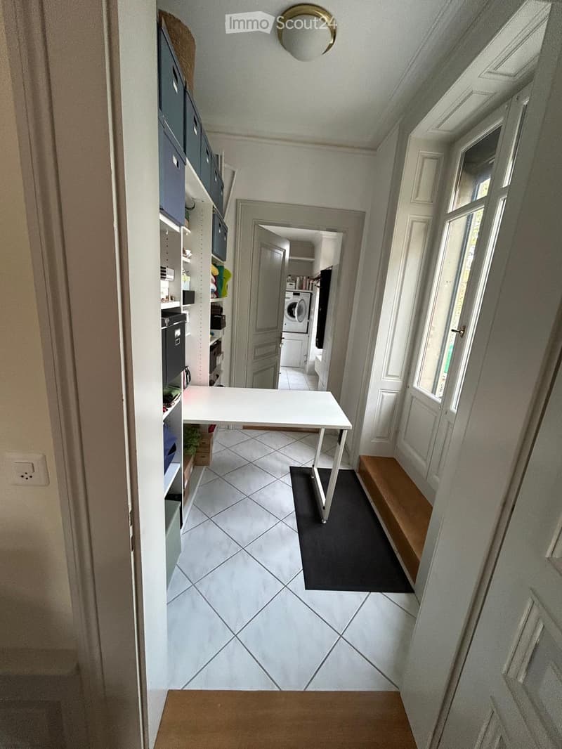 A narrow hallway with tiled flooring, a small white table, a wooden bench, and a shelf with storage boxes. A door leads to a room with a washing machine visible.