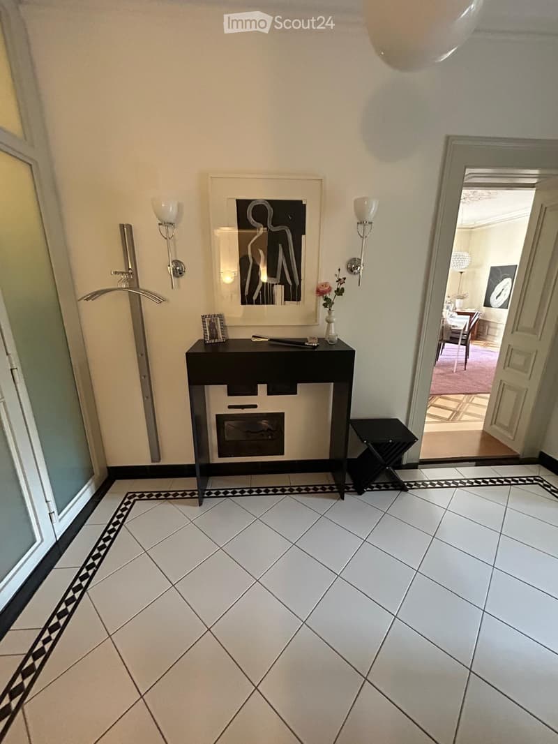 Hallway with white tiled floor, black and white border design, wall-mounted lamps, a black console table with a vase and picture frame, and an open doorway leading to another room.