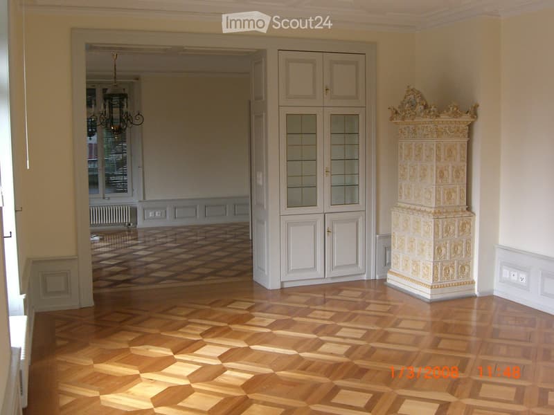 Elegant hallway with polished wooden flooring, ornate decorative column, white cabinet with glass doors, and chandelier visible in the adjacent room.