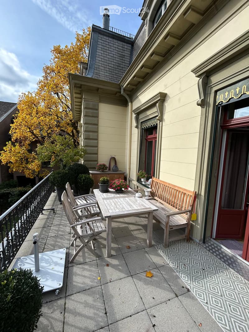 Balcony with a wooden table, bench, and chairs, surrounded by potted plants and a decorative railing.