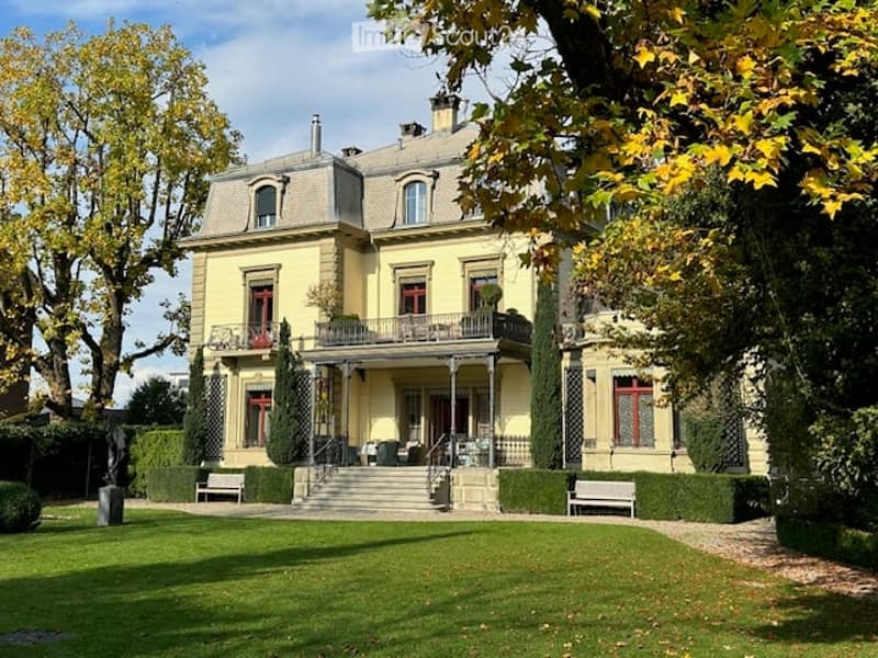 A large, elegant yellow mansion with multiple stories, grey roof, and ornate architectural details. The house features a balcony, manicured lawn, and surrounding trees.