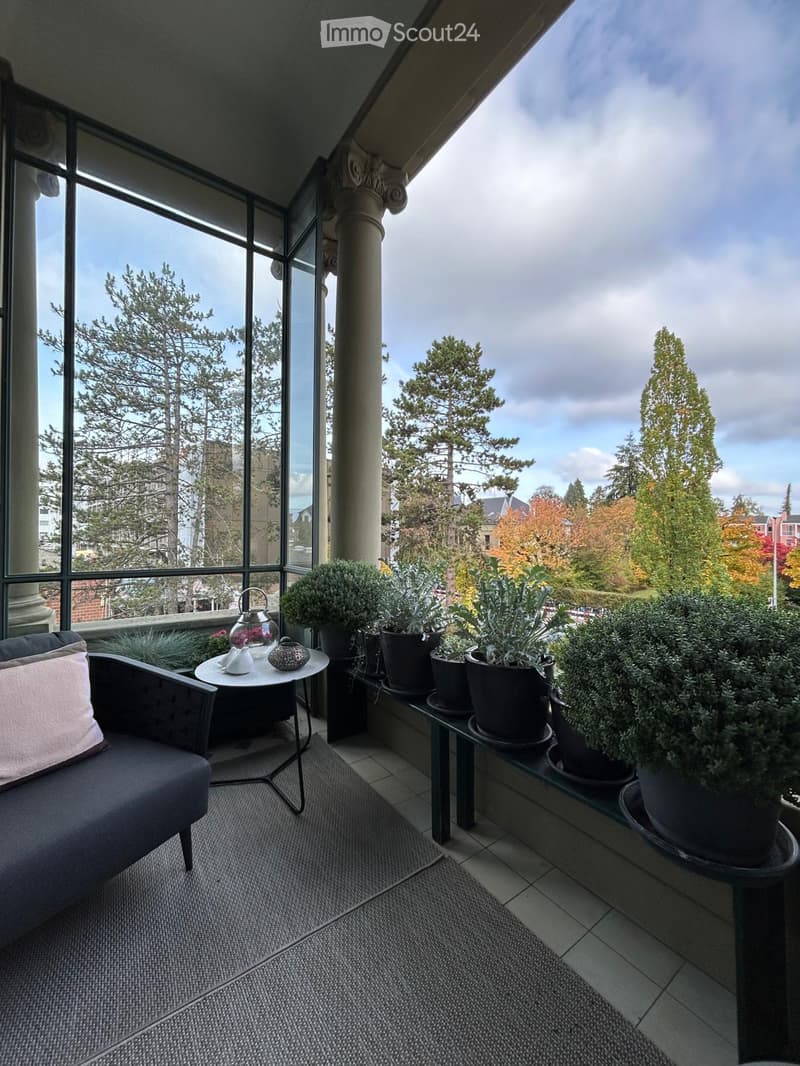 Balcony with large windows, view of trees and cityscape, sofa, small table with teapot, and several potted plants.