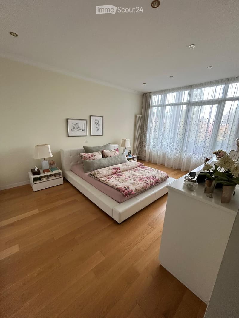 Modern bedroom with wooden flooring, white bed with floral bedding, nightstand with lamp, picture frames on wall, and a small table with flower vase.