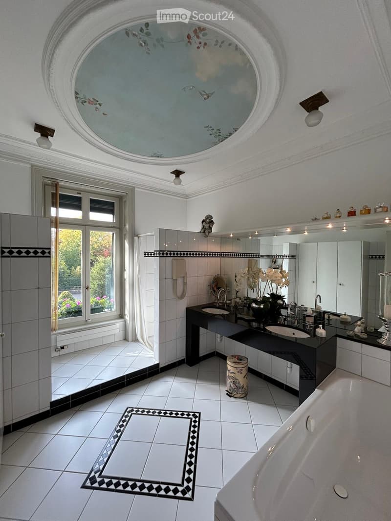 Elegant bathroom with a large bathtub, double sink vanity, decorative mirror, and floral arrangement. Features a round skylight with a painted mural, white tiles, and black accents.