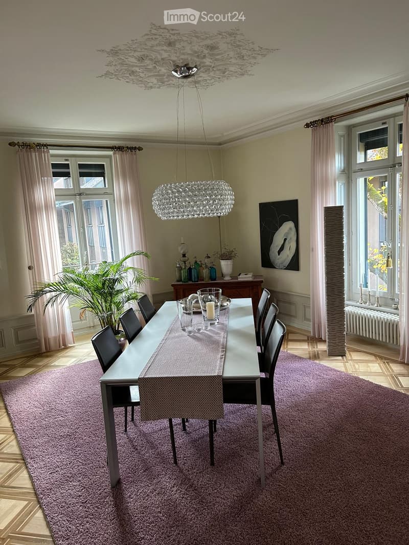 Elegant dining room with a long table set for six, modern chandelier, large windows with curtains, and a decorative painting on the wall.