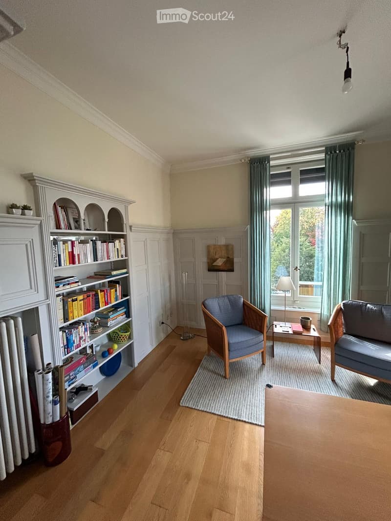 Living room with wooden flooring, large window with curtains, bookshelf filled with books, sofa, armchair, small table, and a lamp.