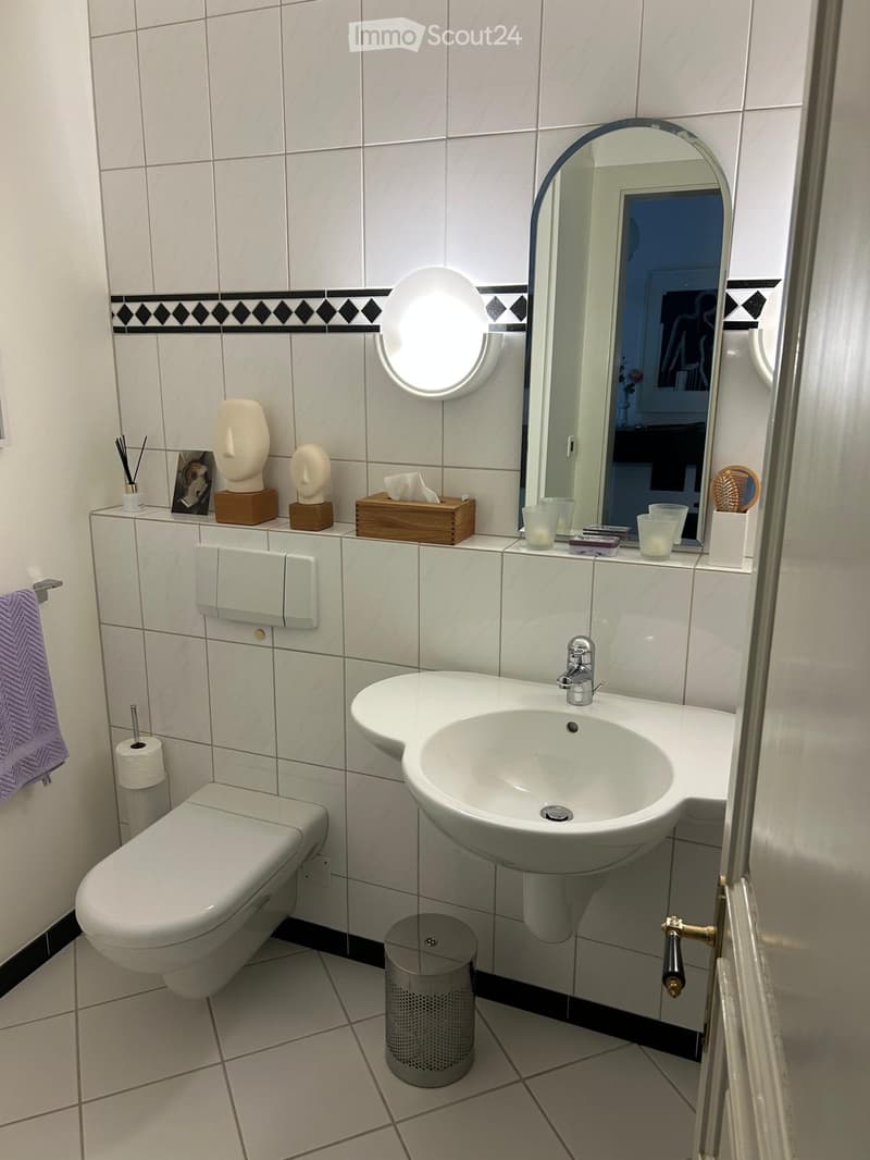 Modern bathroom with white tiles, a sink, toilet, mirror, and decorative items on the counter.