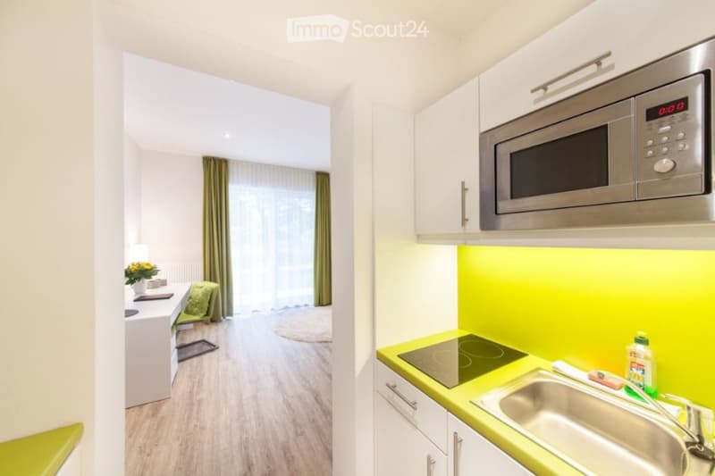 A modern, compact studio apartment with a small kitchen area featuring a microwave, sink, and faucet, and a living space with a chair, table, and a small round rug.