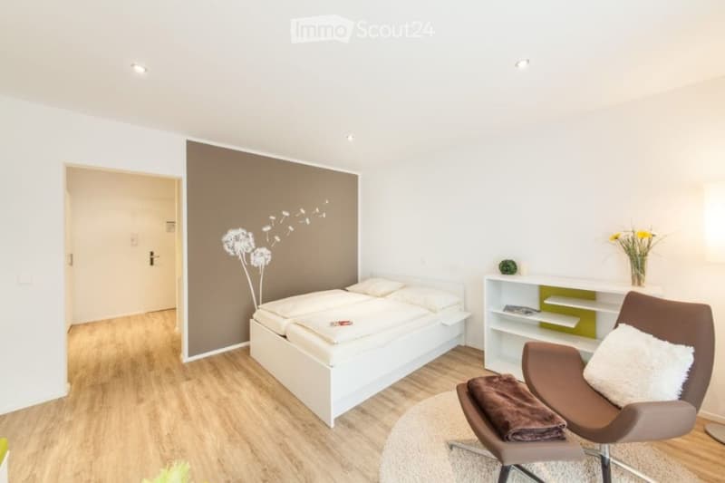 Modern bedroom with a white bed, a brown armchair, a white shelf, and a vase with flowers on the shelf.