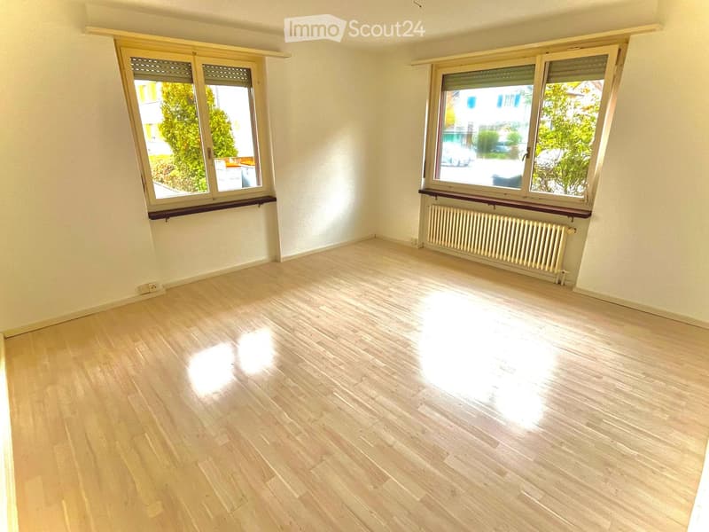 Empty room with light wooden flooring, white walls, and large windows with wooden sills. Radiator visible under one window.