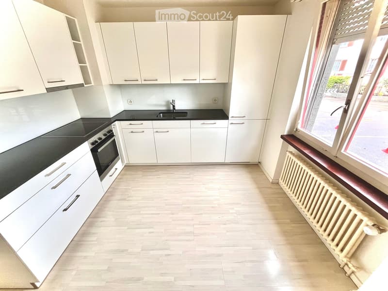 Modern kitchen with white cabinets, black countertops, and a built-in oven. Features a sink with a faucet, a radiator under the window, and light wooden flooring.