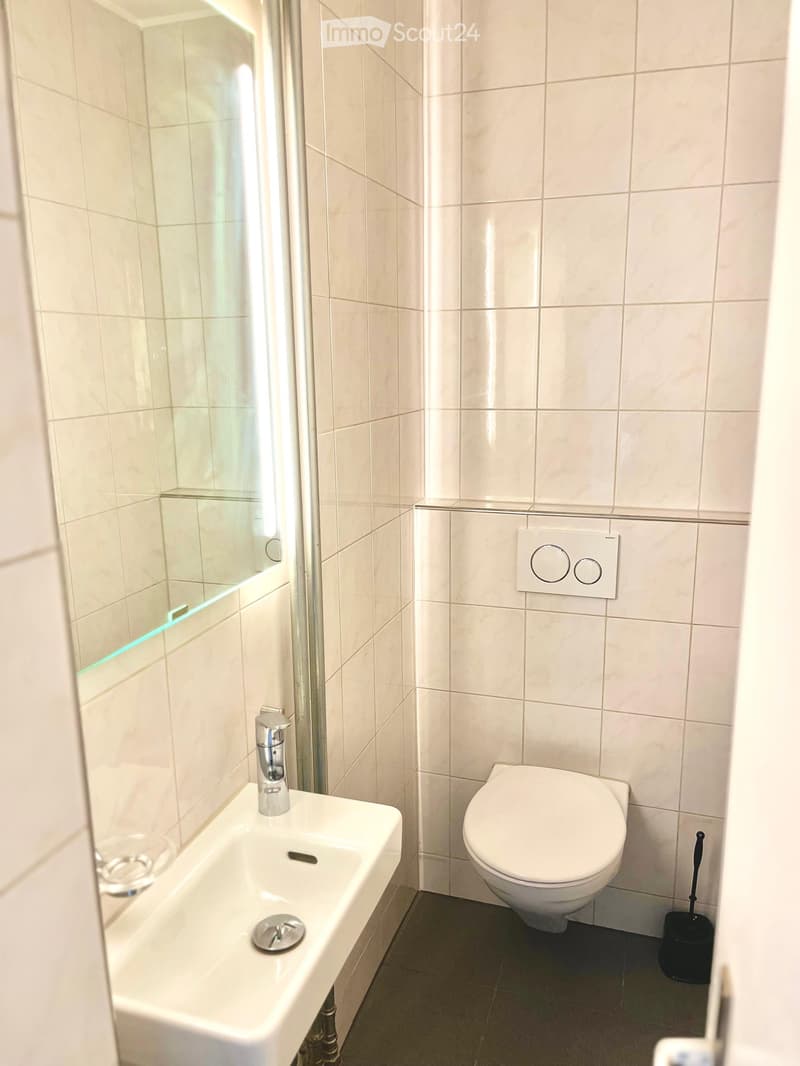 Modern bathroom with white tiles, a white sink with a chrome faucet, a toilet, a mirror, and a light switch on the wall.