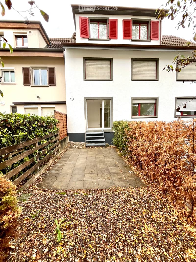 Two-story house with white exterior, red shutters, and a tiled roof. The entrance features a small staircase leading to a door, surrounded by a garden with shrubs and a wooden fence.
