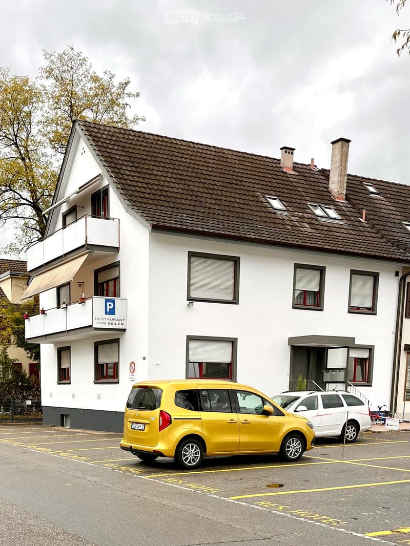 Two-story building with a brown tiled roof, white walls, multiple windows, and a balcony on the left side. A yellow car is parked in front of the building.