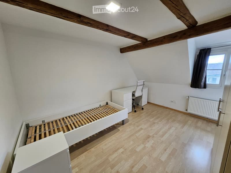 Cozy attic bedroom with wooden slatted bed, white desk, and chair. Exposed wooden beams on the ceiling, wooden flooring, and a window with dark curtains.