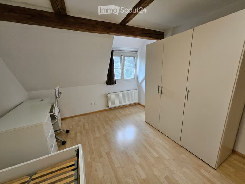 Spacious attic room with wooden floors, exposed wooden beams, large window with curtain, and built-in wardrobe.