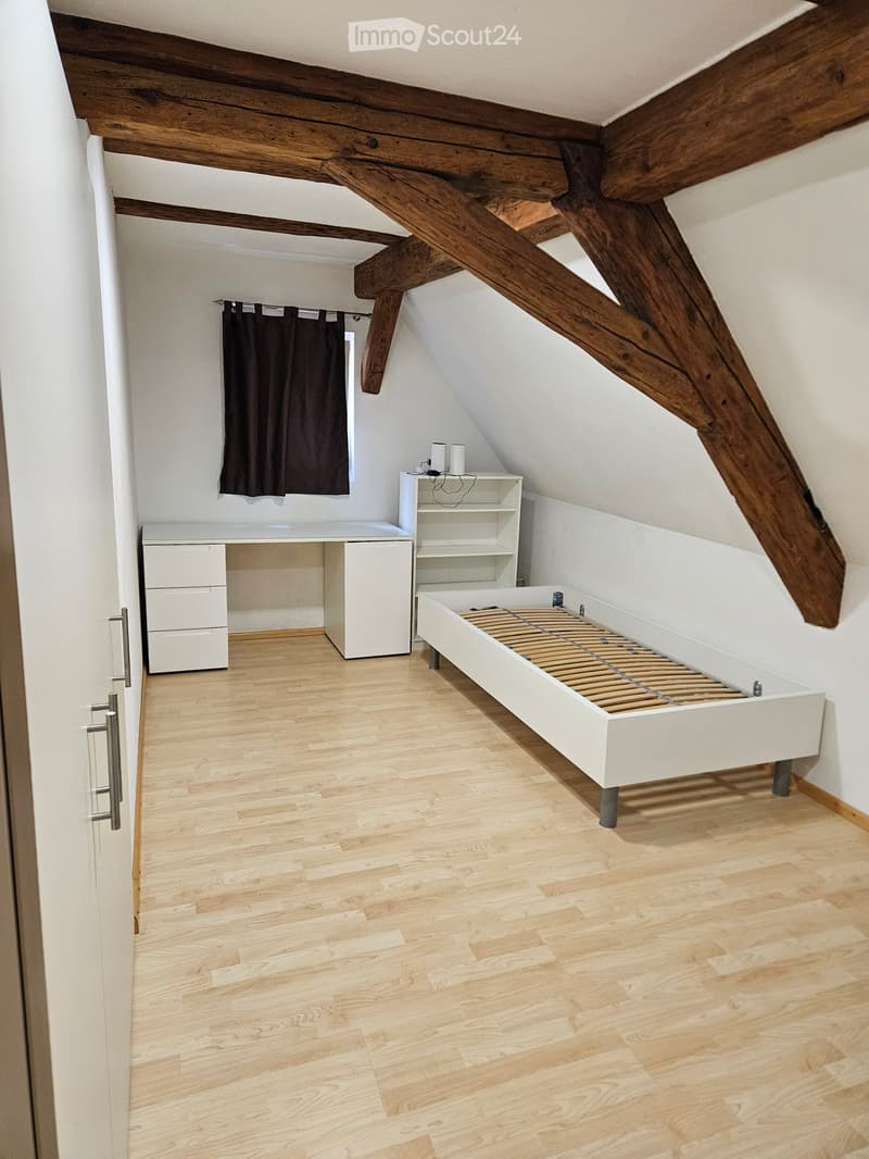 Attic bedroom with wooden beams, white desk, white cabinet, shelf, and a bed with a slatted base.