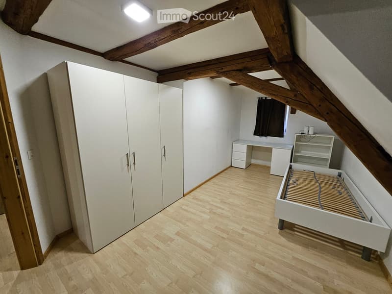 Spacious attic bedroom with wooden beams, light wooden flooring, white wardrobe, desk, shelf, and a bed frame.