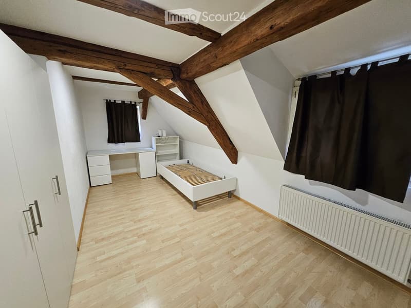 Cozy attic bedroom with wooden beams, light wooden flooring, a single bed, a desk, and a shelf.