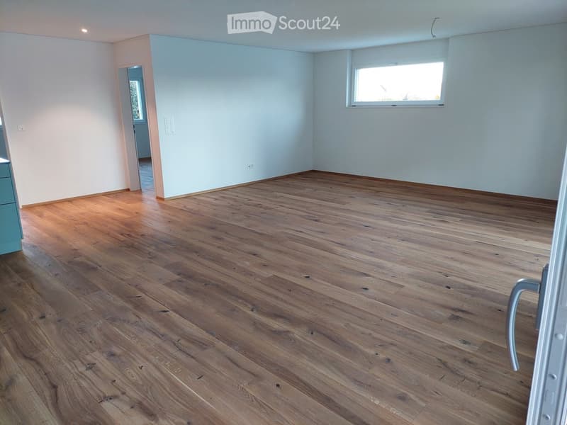 Empty room with wooden flooring, light blue walls, and a large window letting in natural light. The room has a door leading to another space.