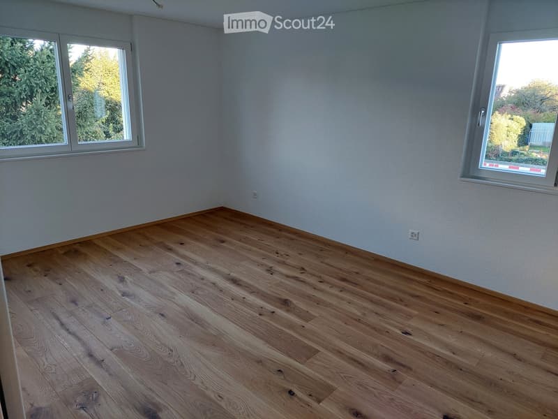 Empty room with light wooden flooring and large windows offering a view of greenery outside.