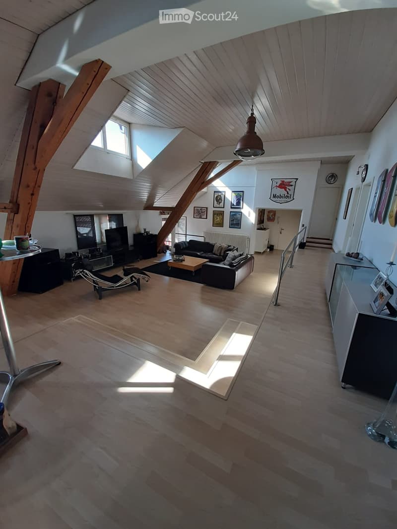 Bright, open living space with wooden floors, exposed wooden beams, and skylights. Features a black sectional sofa, coffee table, modern chair, TV, and decorative picture frames.