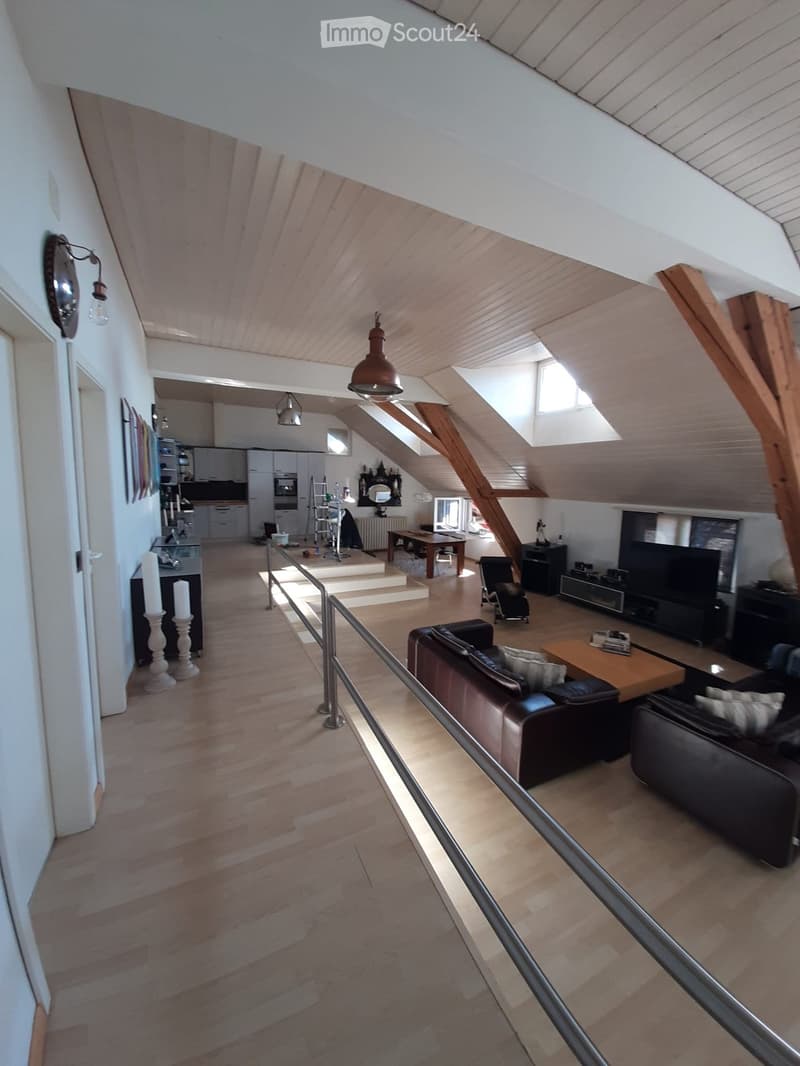Open-plan living and dining area with wooden flooring, exposed wooden beams, and skylights. Features a leather sofa, coffee table, TV, dining table, and kitchen area with cabinets and appliances.