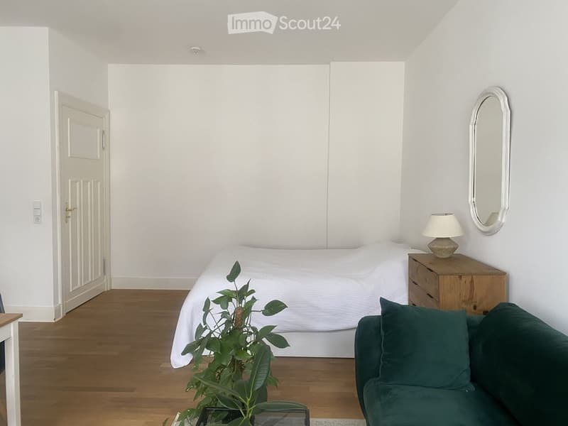 A small, modern bedroom with a bed, green sofa, nightstand with a lamp, and a decorative mirror on the wall. The room features wooden flooring and white walls.