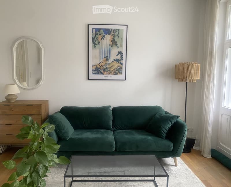 Modern living room with a green velvet sofa, glass coffee table, floor lamp, table lamp, mirror, plant, and artwork on the wall.