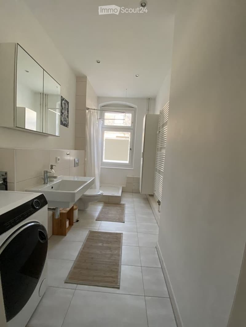 Modern bathroom with white tiles, a sink, toilet, shower, and a washing machine. Features a large mirror and two bath mats on the floor.