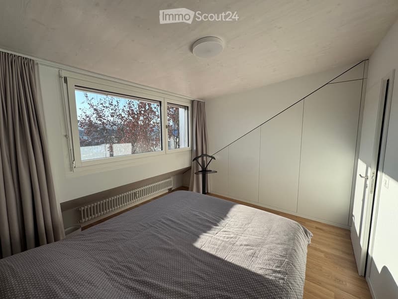 Modern bedroom with a bed, built-in wardrobe, large window with curtains, radiator, and a clothes hanger stand.