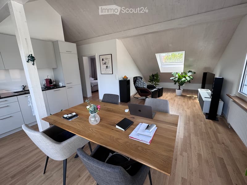 A modern, open-plan living and working space with a wooden table, laptop, chairs, kitchen area, and a comfortable seating area with a plant and screen.