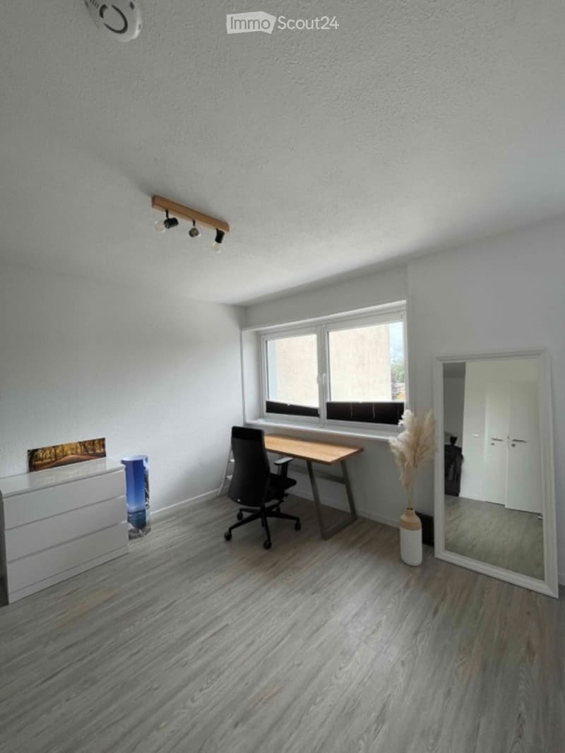 Modern, minimalist room with light grey walls, wooden floor, large window, desk, chair, mirror, and decorative vase.