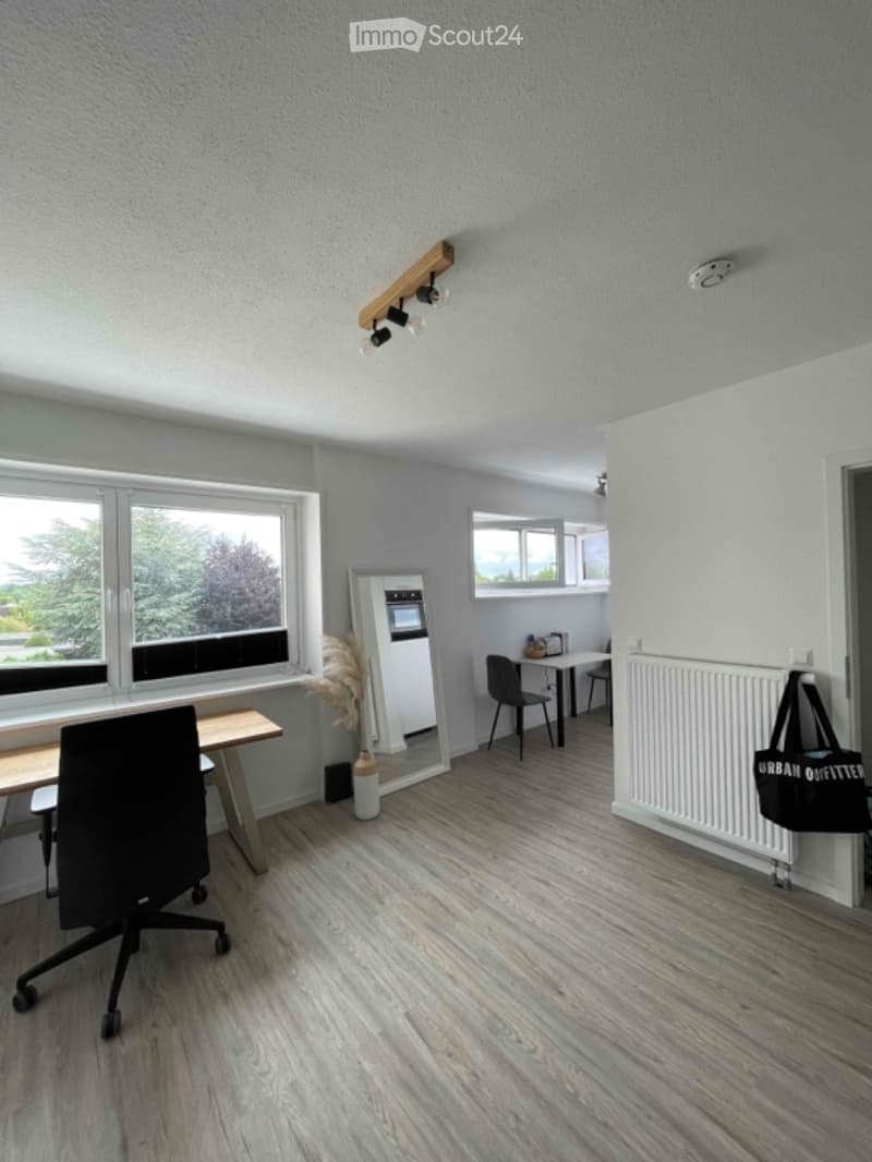 A modern, well-lit room with light grey walls and wooden flooring. It features a large window with a view, a small kitchen area with a refrigerator, and a workspace with a chair and table. A radiator is mounted on the wall, and a black bag hangs nearby.