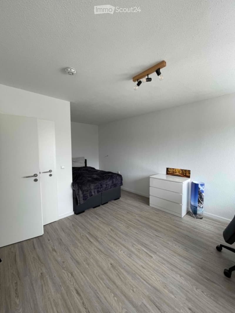 Modern bedroom with light grey walls, wooden flooring, a bed with dark bedding, a white nightstand, and a chair near the door.