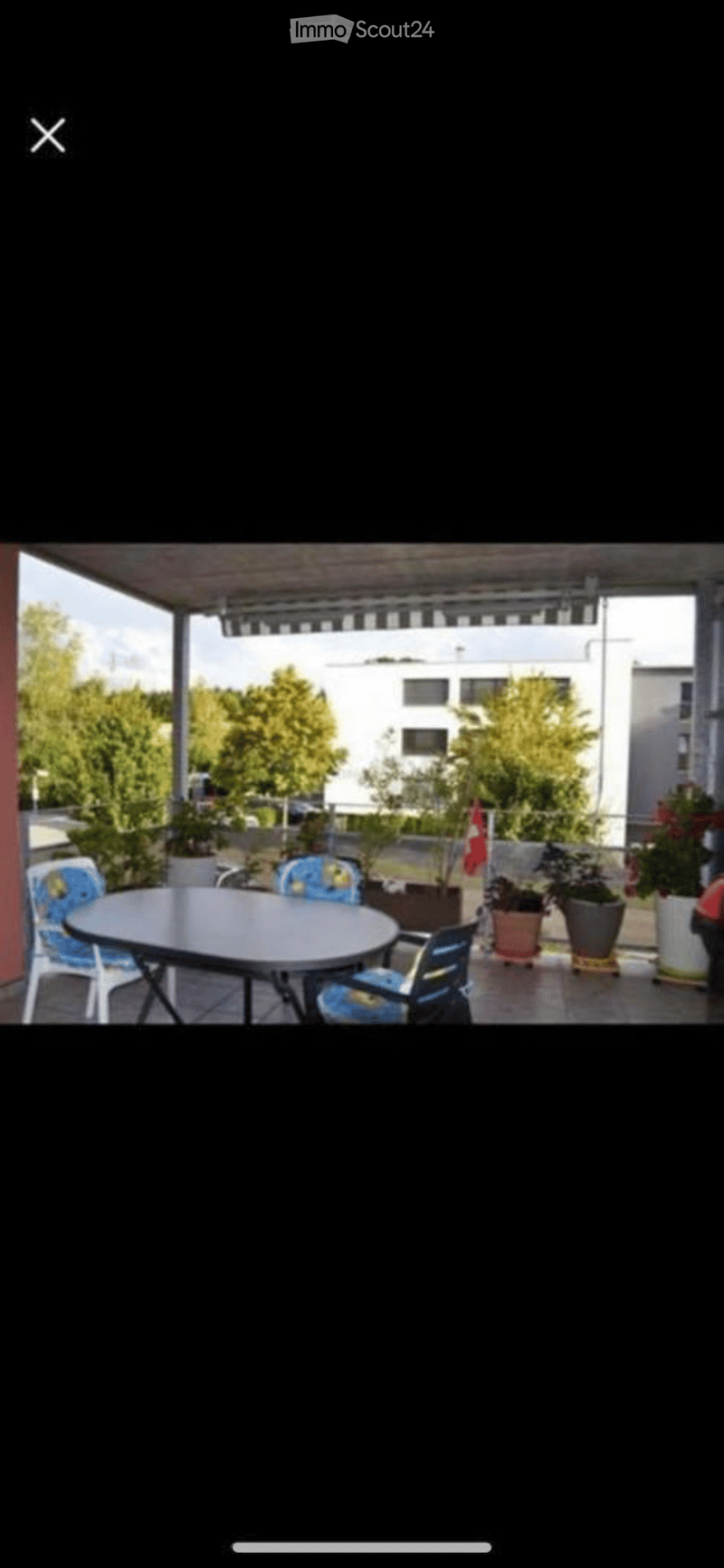 Balcony with a round table and chairs, surrounded by potted plants and trees, with a covered roof and a view of the building and greenery.