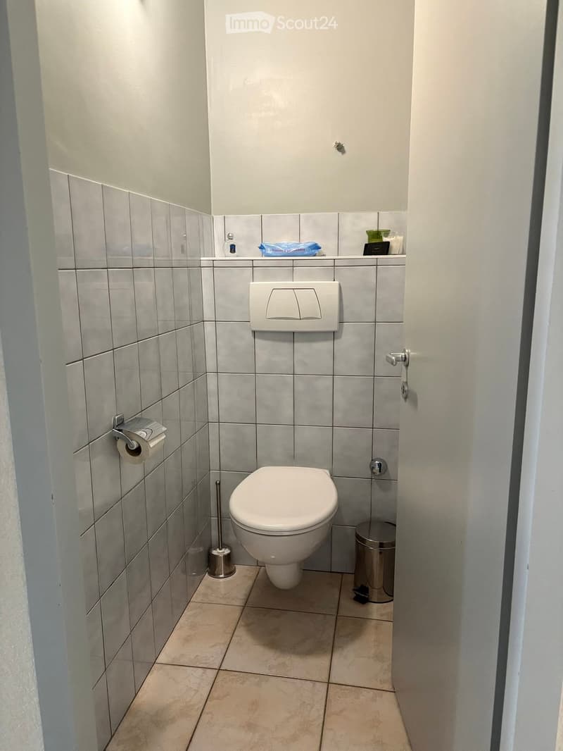 Modern bathroom with grey tiled walls, white toilet, toilet paper holder, and a small trash bin. The floor is tiled in a light beige color.