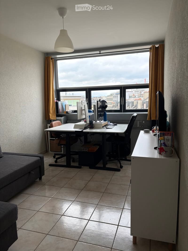 A modern, well-lit room with a large window offering a city view. It features a sofa, a desk with a computer setup, two chairs, and a lamp hanging from the ceiling.