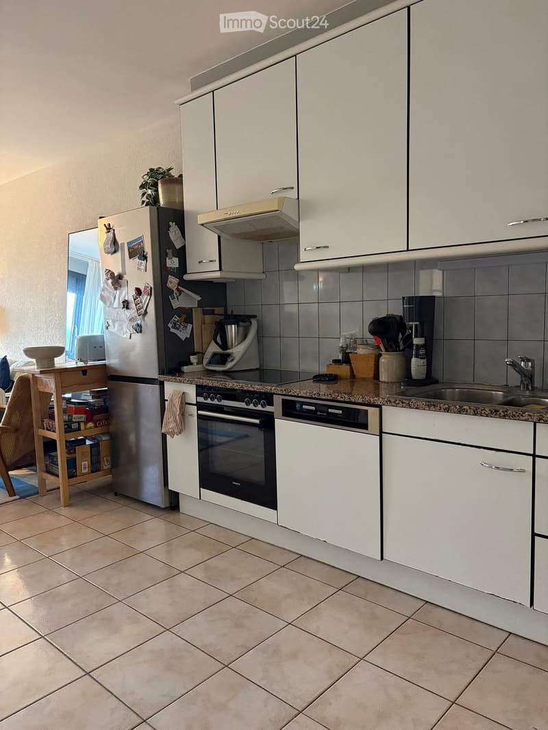 Modern kitchen with white cabinets, granite countertop, stainless steel appliances including a refrigerator, oven, dishwasher, and sink. A small wooden stool holds various items, and a bowl is placed on the counter.