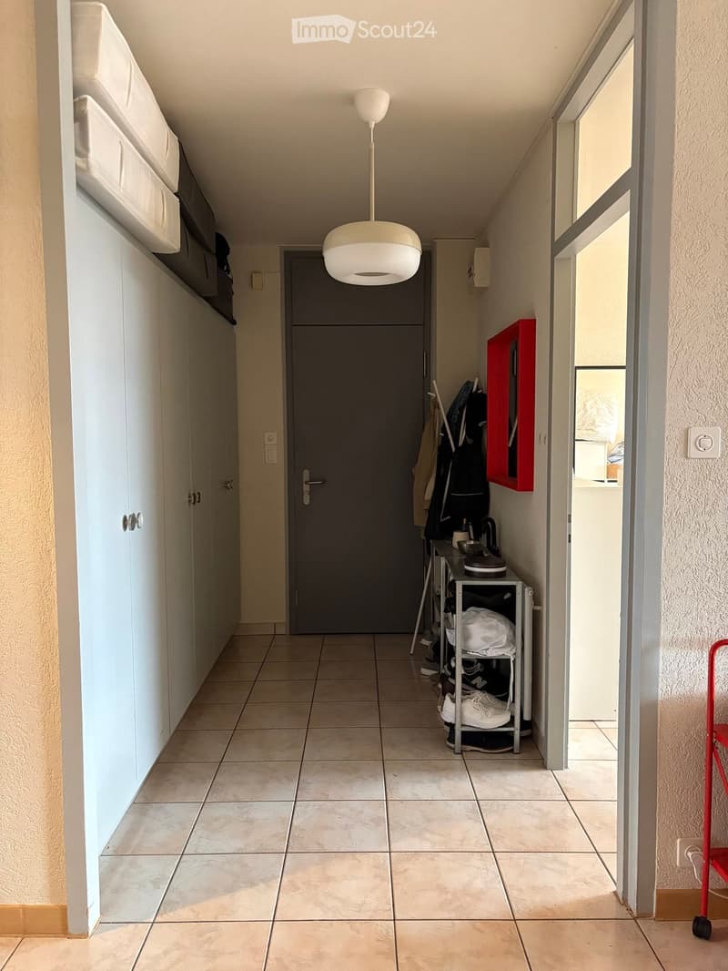 Clean hallway with tiled floor, white walls, built-in storage, and a hanging light fixture. A small shelf holds shoes and bags, and a red storage unit is visible on the right.